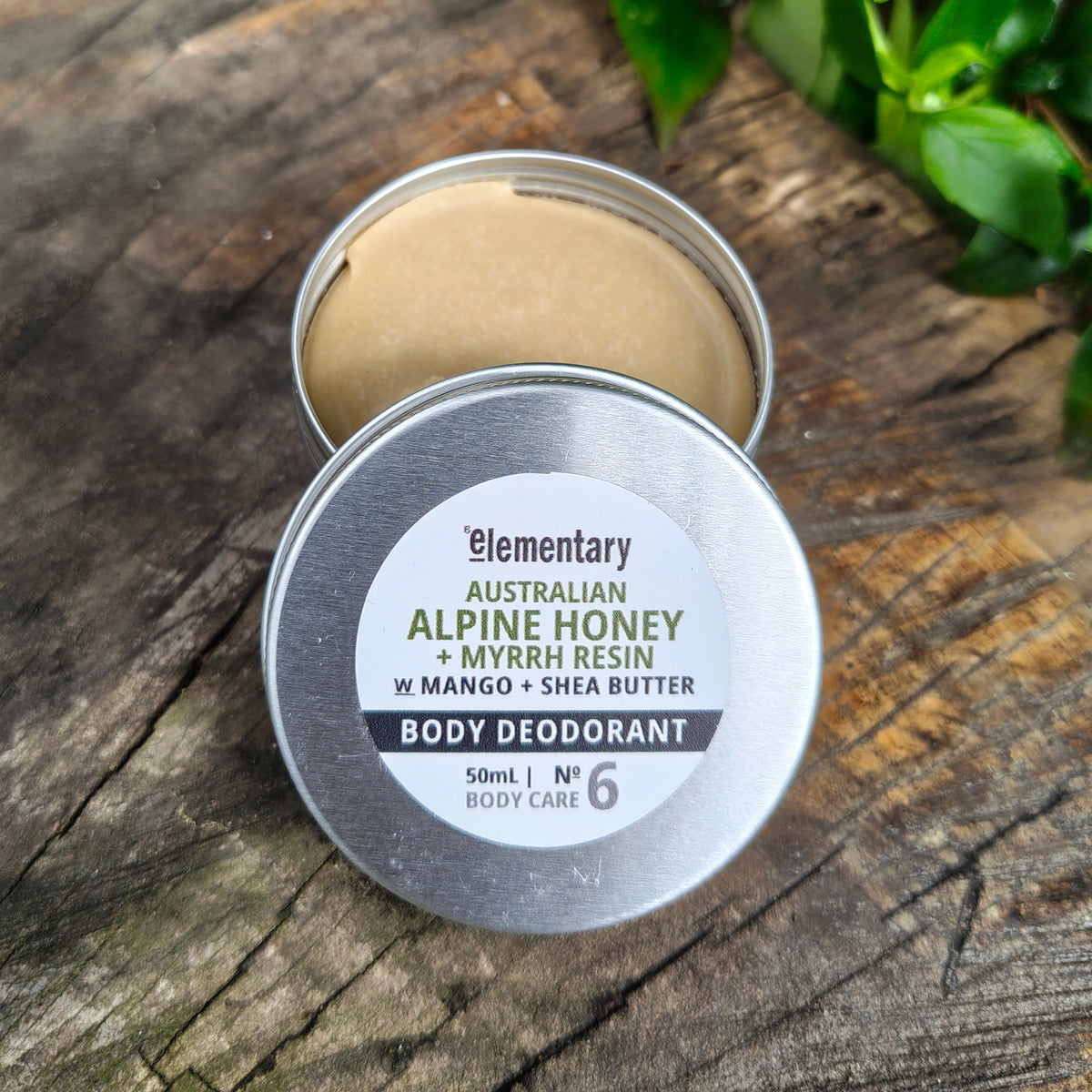 An open round metal tin of body deodorant with the lid placed in front, showing the natural deodorant paste inside. The tin is photographed on a rustic wooden surface with green leaves in the background.