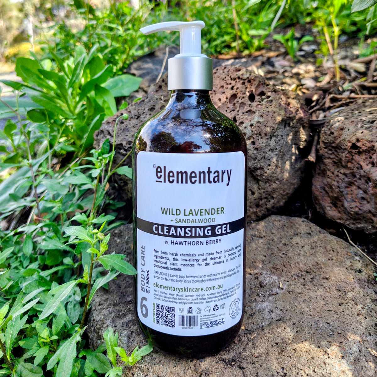 Elementary Wild Lavender Cleansing Gel in a dark amber pump bottle, placed on a natural stone surface surrounded by green plants outdoors.