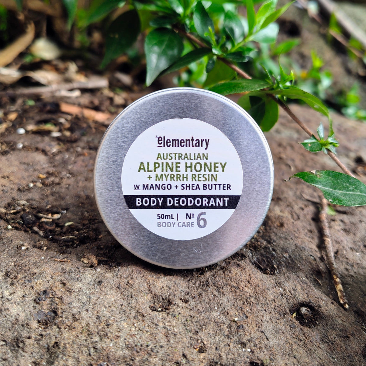 A round silver tin of Elementary Australian Alpine Honey Body Deodorant resting on a textured brown rock with green plants in the background.