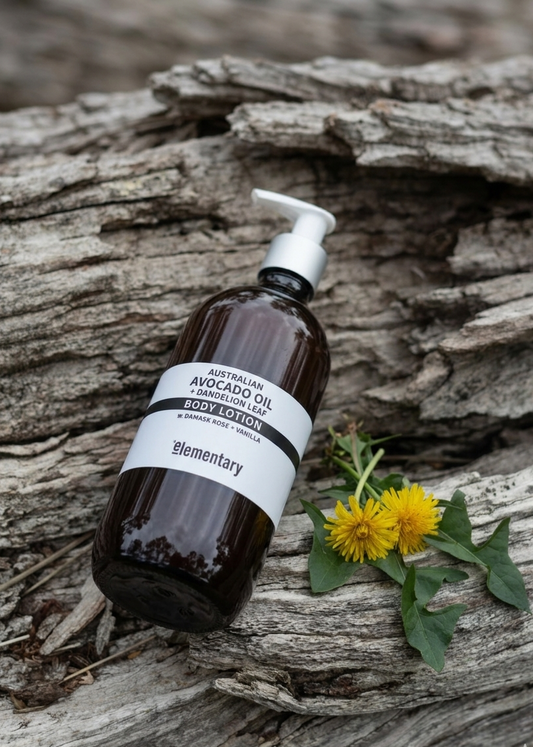 Avocado and Dandelion Leaf Body lotion bottle by Elementary Skin Care with on a driftwood surface with Dandelion flowers.