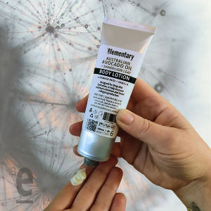A hand holding a squeeze tube of Elementary Australian Avocado Oil Body Lotion, with another hand near the bottom of the tube, set against a white background with a faint dandelion illustration.