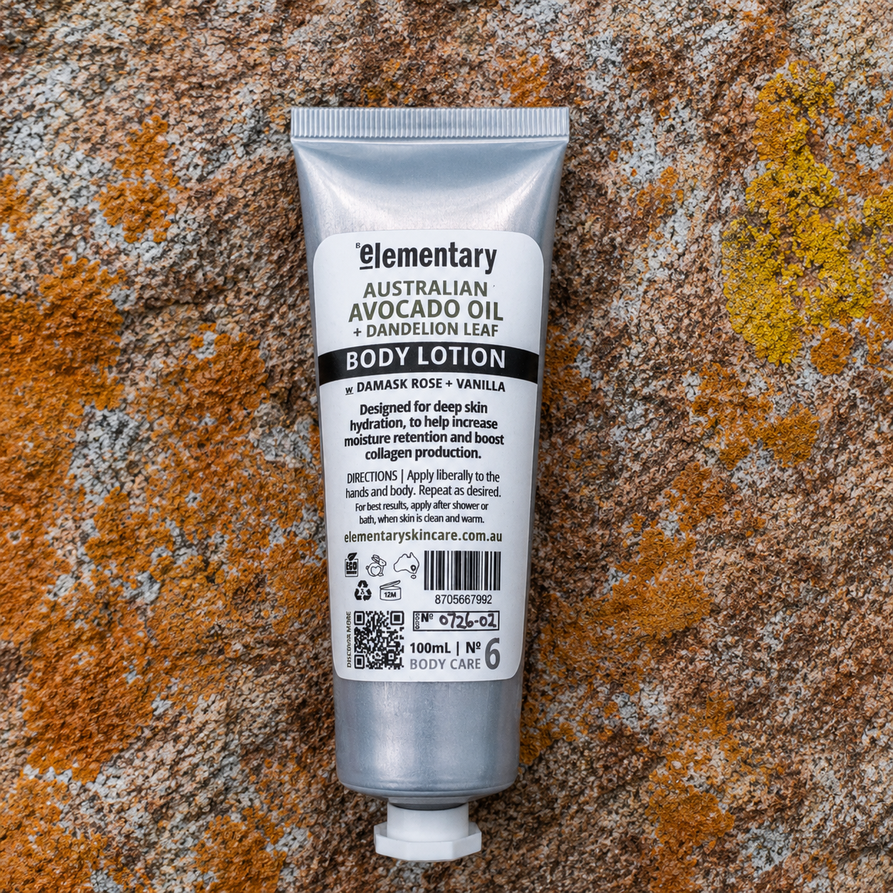 A 100ml squeeze tube of Elementary Australian Avocado Oil + Dandelion Leaf Body Lotion, in Damask Rose + Vanilla scent, placed flat on a textured, orange and brown lichen-covered rock surface.