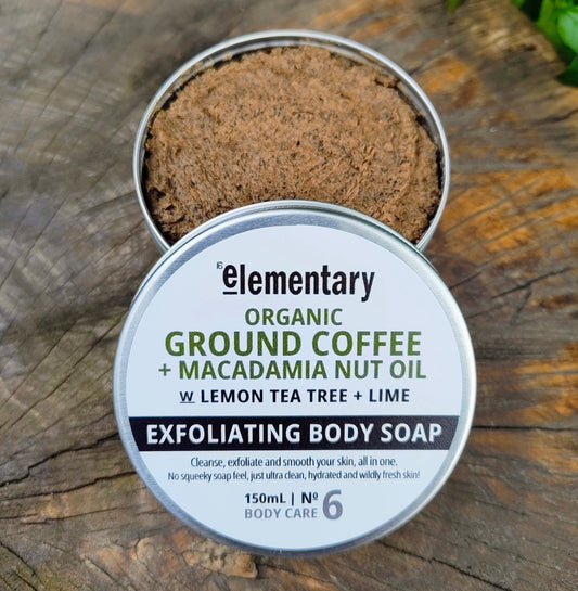 An open round metal tin of Elementary Exfoliating Body Soap, showing the brown exfoliating ground coffee and macadamia nut oil inside, placed on a rustic wooden surface with a branded white lid in front.
