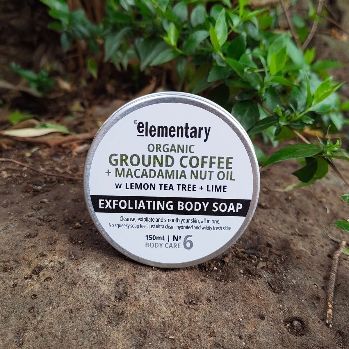 A round tin of Elementary Organic Exfoliating Body Soap placed on a textured stone surface outdoors, with green foliage and brown branches in the background. The label reads 'Elementary Organic Ground Coffee + Macadamia Nut Oil + Lemon Tea Tree + Lime'.