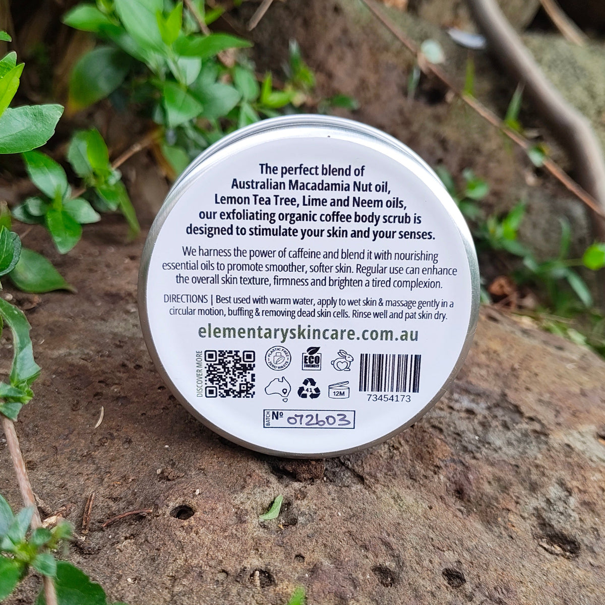 A round tin of Elementary Skin Care 'Macadamia & Coffee Body Scrub' placed on a natural rock surrounded by green leaves and plants outdoors.