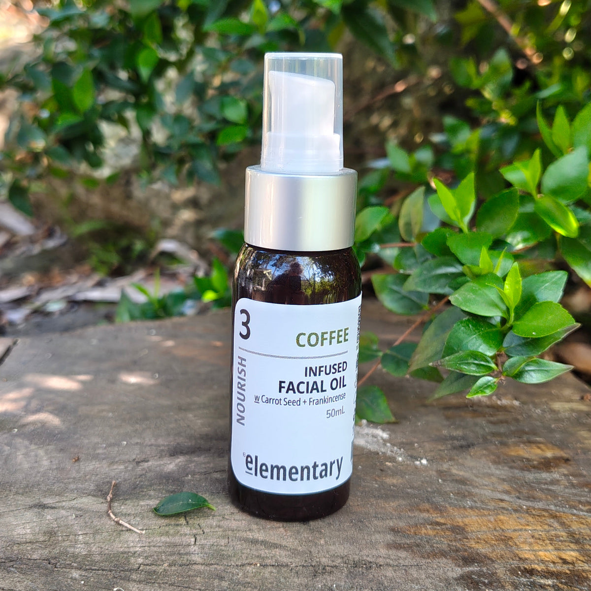 A 50ml bottle of Elementary Coffee Infused Facial Oil, featuring a dark amber glass bottle, silver pump collar, and white label, set on a rustic wooden surface with a blurred green garden background.