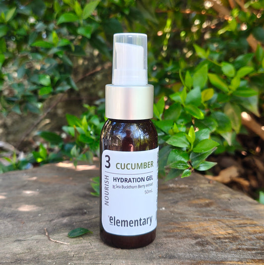 A small amber glass spray bottle labeled 'Cucumber Hydration Gel' by the brand elementary, placed on a weathered wooden surface with a blurred background of green foliage.