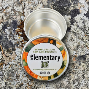 An open round metal tin and its lid placed on a textured stone surface. The lid label reads 'Earth Conscious Skin Care Products' and 'elementary' with a colorful botanical design.