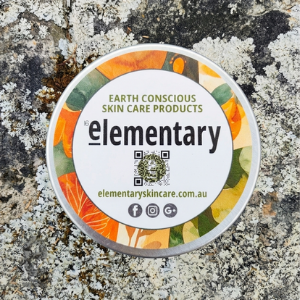 A round tin of Elementary Earth Conscious Skin Care Products, featuring a white label with orange and green botanical illustrations, placed on a textured gray and white lichen-covered rock surface.