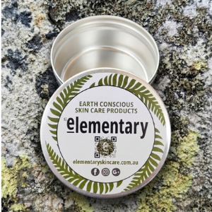 An open round metal tin of skin care product with its lid placed in front, showing a white label with green fern leaves and the brand name 'elementary'. The tin and lid are photographed on a textured gray and white stone surface.