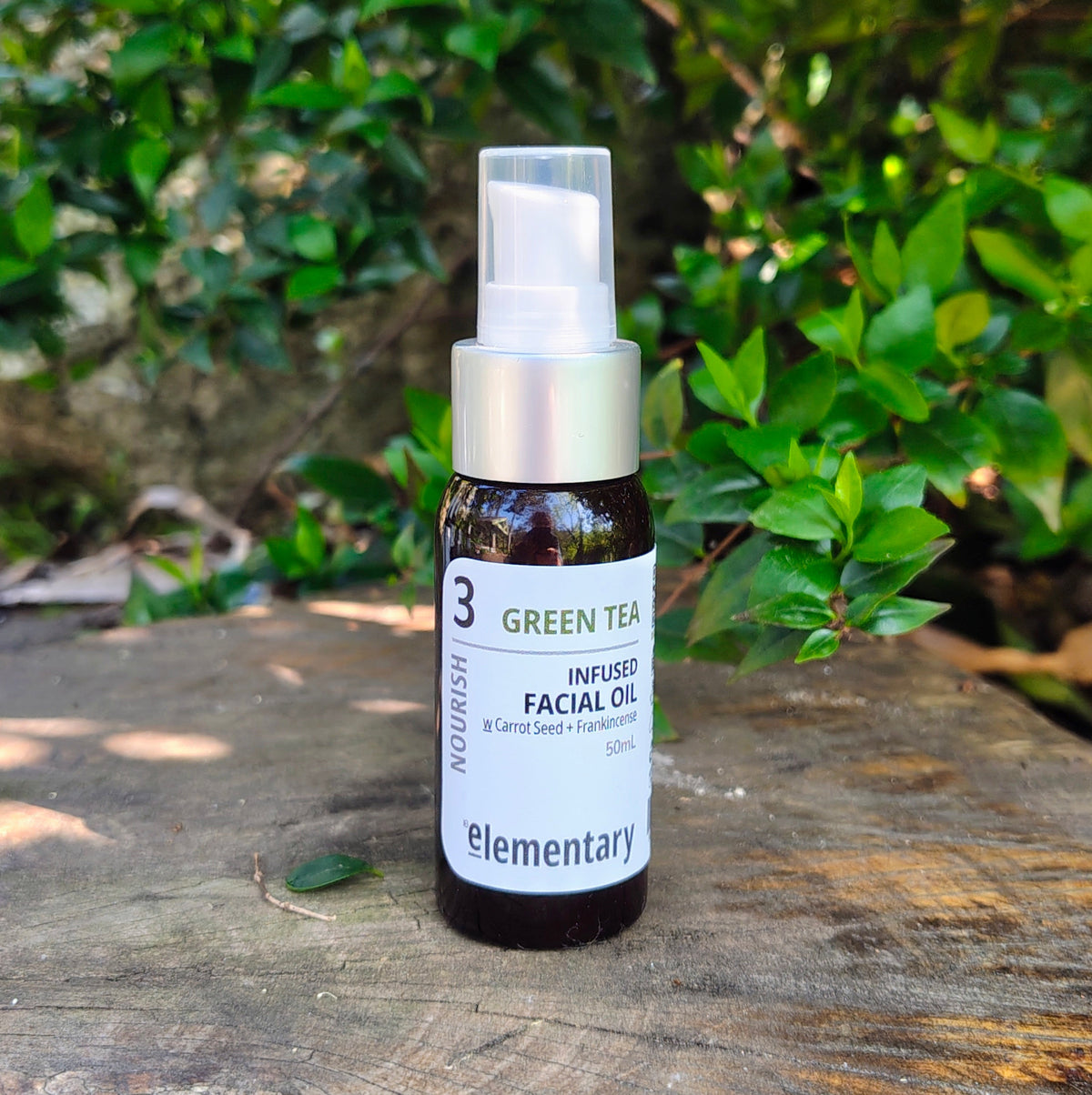 A small amber glass bottle labeled 'elementary nourish green tea infused facial oil' with a white label and silver pump, standing on a weathered wooden surface with green foliage in the background.