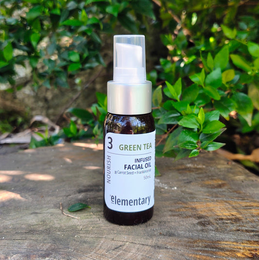 A small amber glass bottle labeled 'elementary nourish green tea infused facial oil' with a white label and silver pump, standing on a weathered wooden surface with green foliage in the background.