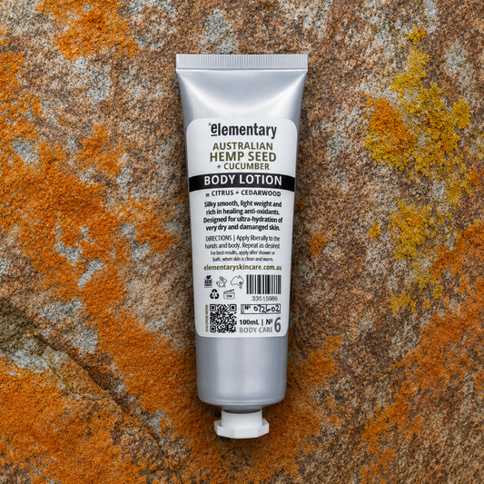 A 100ml squeeze tube of Elementary Australian Hemp Seed + Cucumber Body Lotion. The tube is metallic silver with a white cap, set against a textured, rustic background of orange and gray stone.