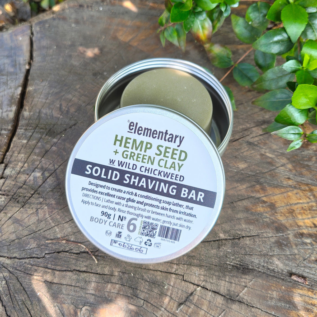 A round metal tin of 'Elementary Hemp Seed + Green Clay' solid shaving bar sits open on a weathered wooden log outdoors, with green leaves in the background.