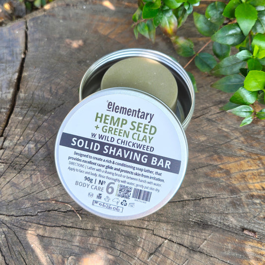 A round metal tin of 'Elementary Hemp Seed + Green Clay' solid shaving bar sits open on a weathered wooden log outdoors, with green leaves in the background.