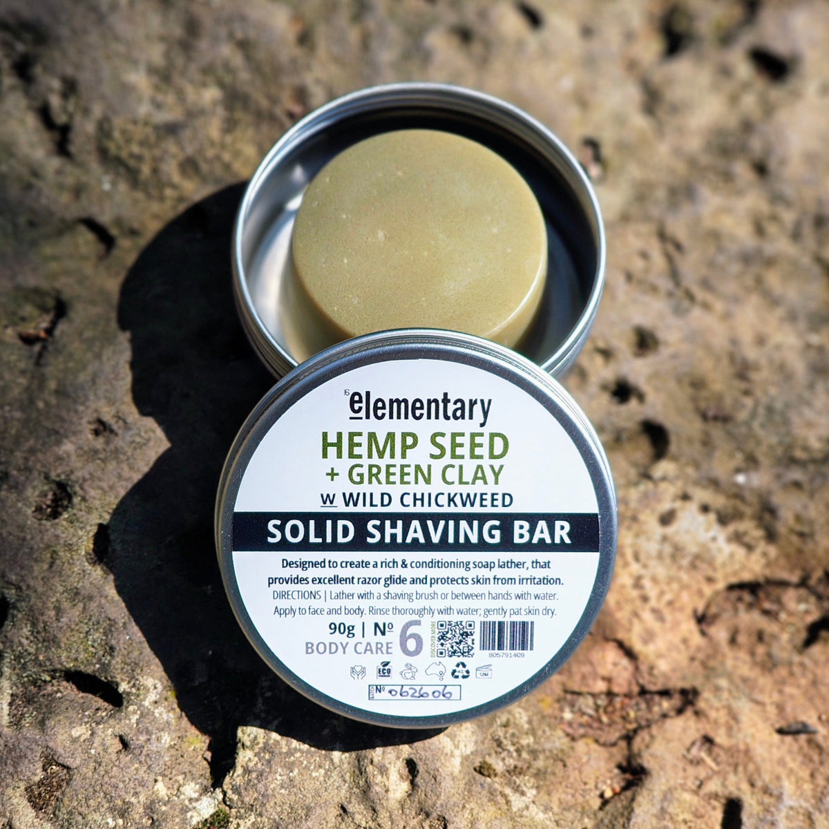 An open round metal tin of Elementary Hemp Seed + Green Clay solid shaving bar sits on a textured stone surface, with the lid placed in front showing the product label. The shaving bar inside is visible and the background is natural rock.
