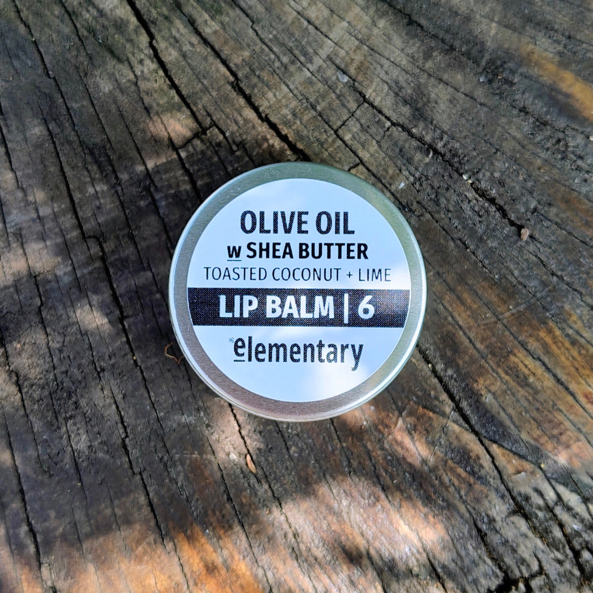 A small round tin of Elementary brand lip balm sits on a weathered wooden surface.