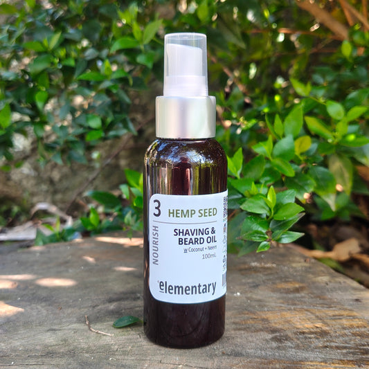 A bottle of Elementary Nourish Hemp Seed Shaving & Beard Oil standing on a weathered wooden surface with a lush green garden background.