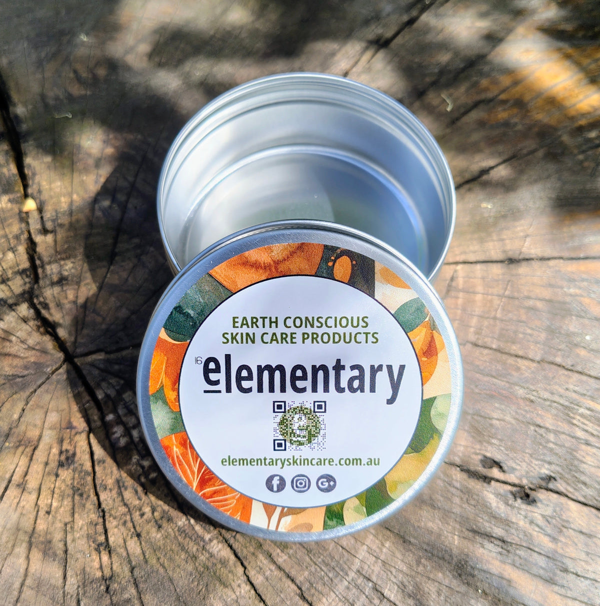An open silver metal tin of 'Elementary' earth conscious skin care products sits on a weathered wooden surface, with the lid placed beside it. The lid features a white label with colorful illustrations of citrus fruits and green leaves.