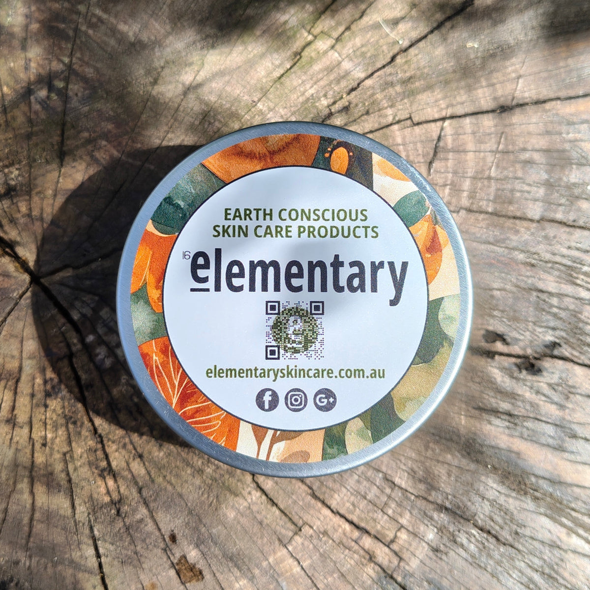 A round tin of Elementary Earth Conscious Skin Care Products placed on a weathered wooden log surface with natural dappled sunlight and shadows.