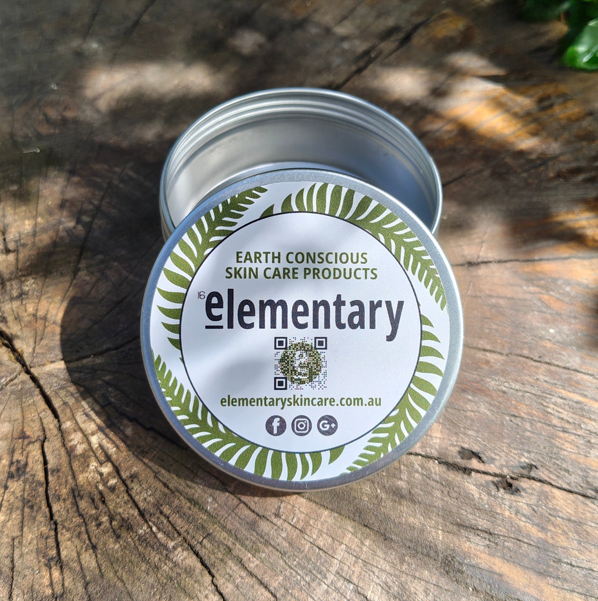An open round aluminium storage tin of skin care product sits on a weathered wooden surface outdoors. The lid label reads 'Elementary' with green leaf illustrations and the website elementaryskincare.com.au