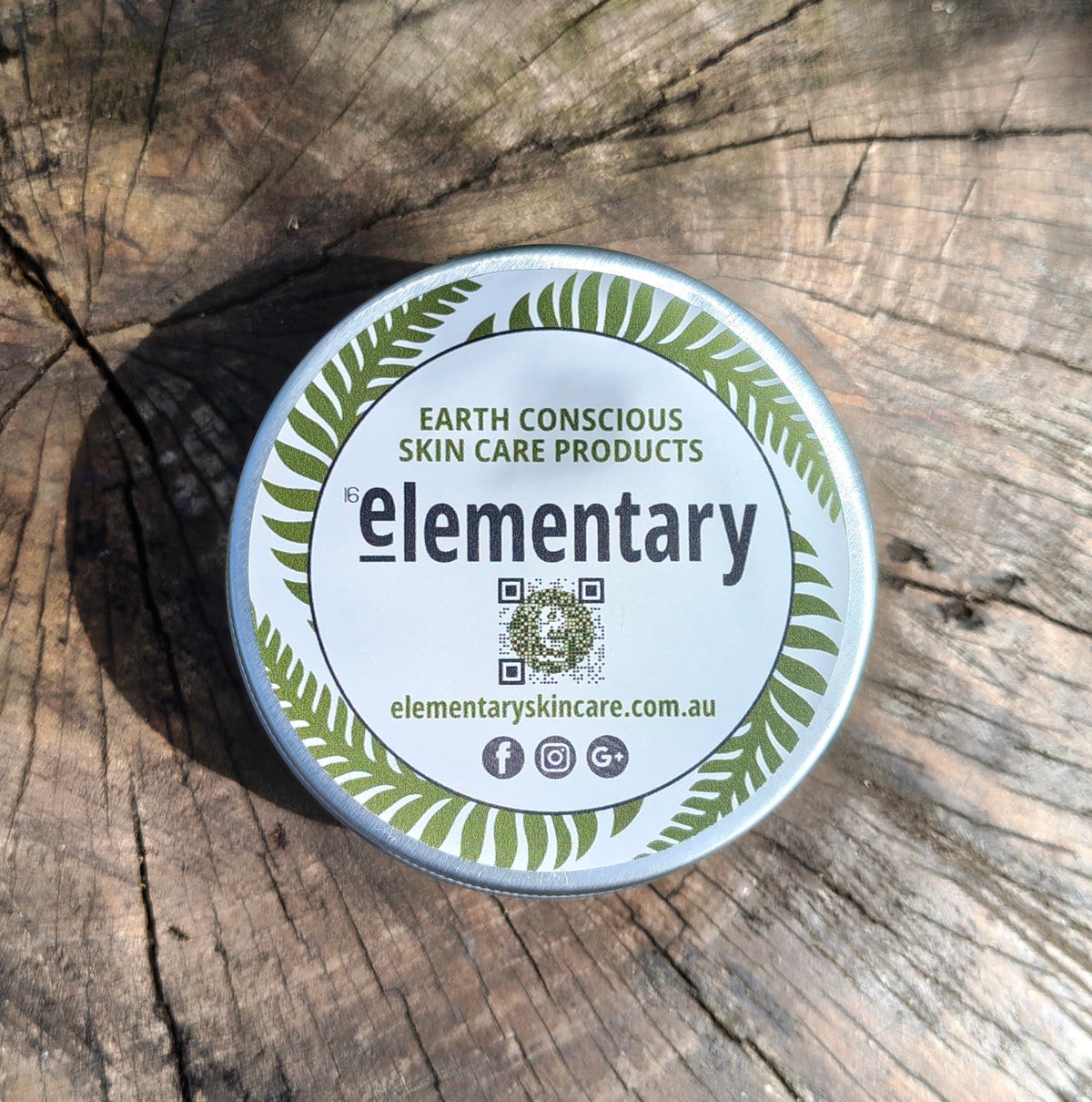 A round metal tin of Elementary Earth Conscious Skin Care Products placed on a weathered wood log surface.