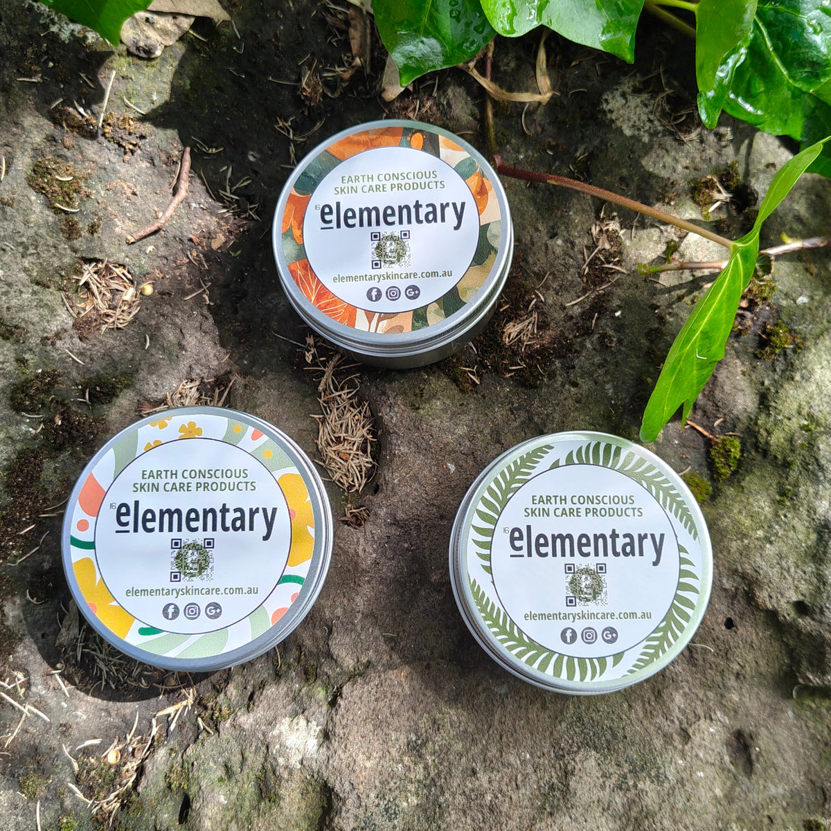 Three round Elementary aluminium storage tins, arranged on a natural stone surface with green leaves in the background.