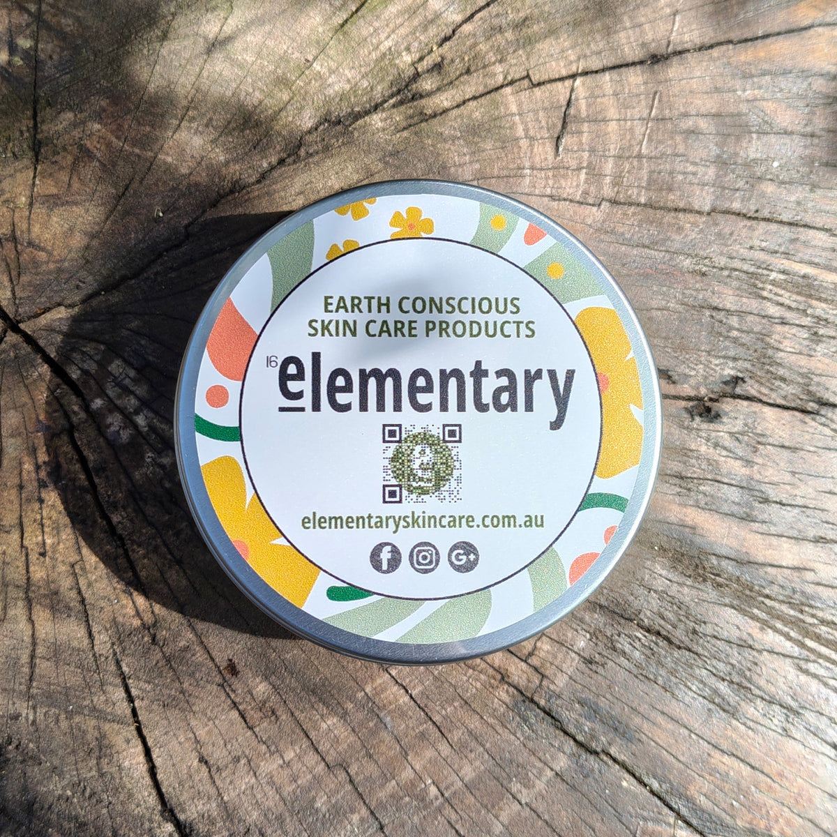 A round tin of Elementary Earth Conscious Skin Care Products sits on a weathered wood log. The tin features a colorful label with yellow, orange, green, and blue abstract shapes on a white background, with the brand name 'elementary' in black text.