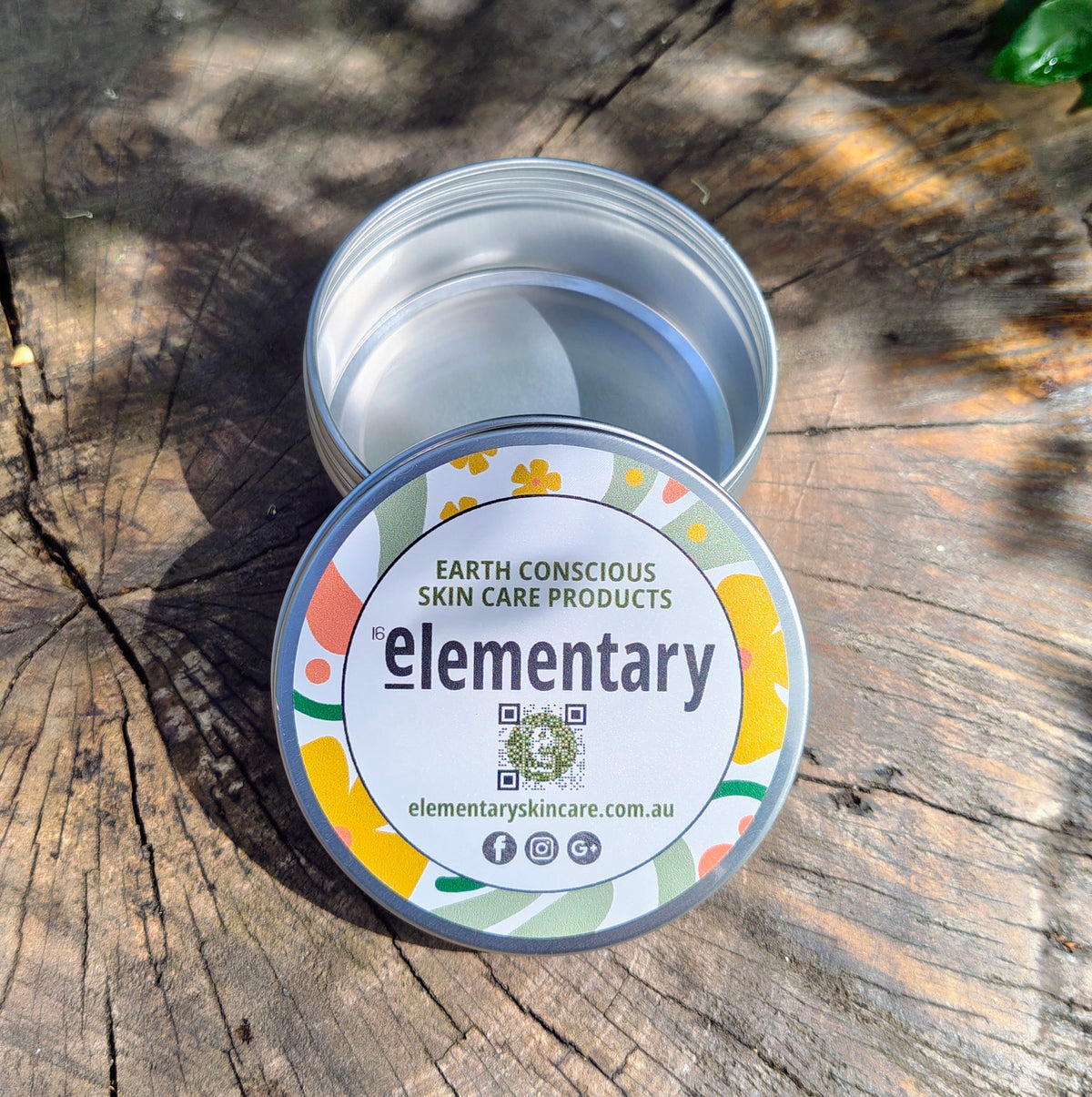 An open round metal tin of skin care product sits on a weathered wooden surface, with the lid placed in front. The lid label reads 'elementary' and 'Earth Conscious Skin Care Products.