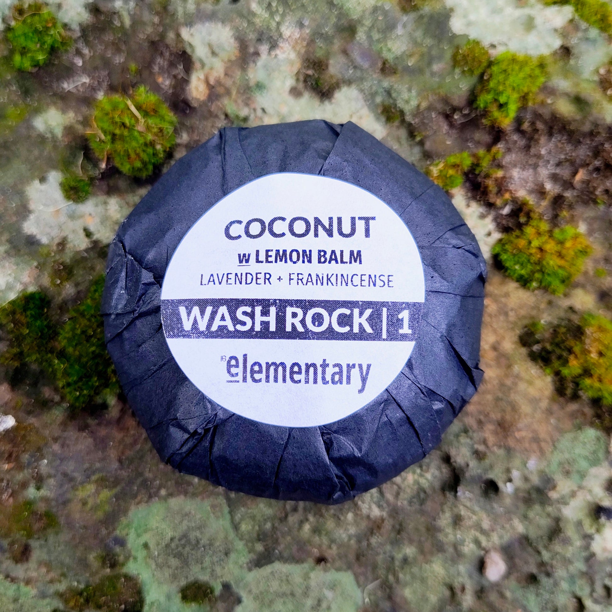 A round wrapped soap bar, COCONUT w/ LEMON BALM' by Elementary SKin Care sits on a mossy rock surface. The soap is wrapped in dark paper with a white circular label.