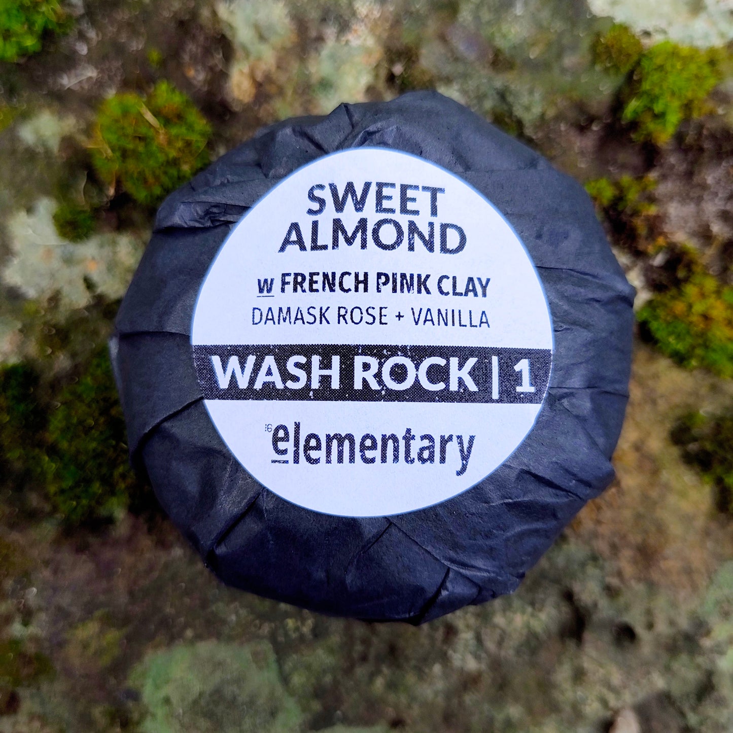 A wrapped Wash Rock by the brand Elementary, labeled 'Sweet Almond w/ French Pink Clay Damask Rose + Vanilla'  placed on a mossy, rocky surface.