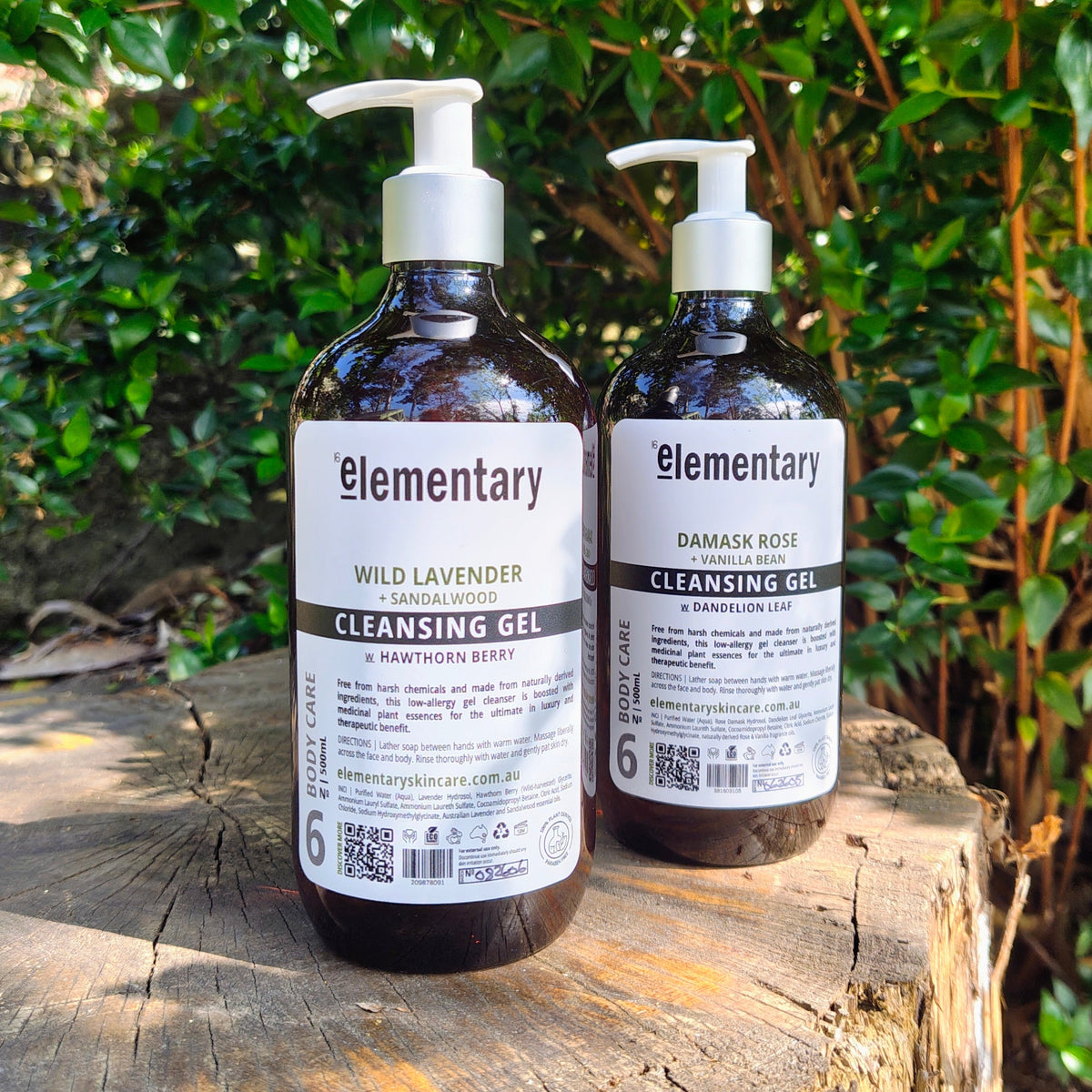 Two dark amber pump bottles of Elementary Cleansing Gel, one labeled Wild Lavender and the other Damask Rose, displayed on a rustic wooden log with a lush green garden background.