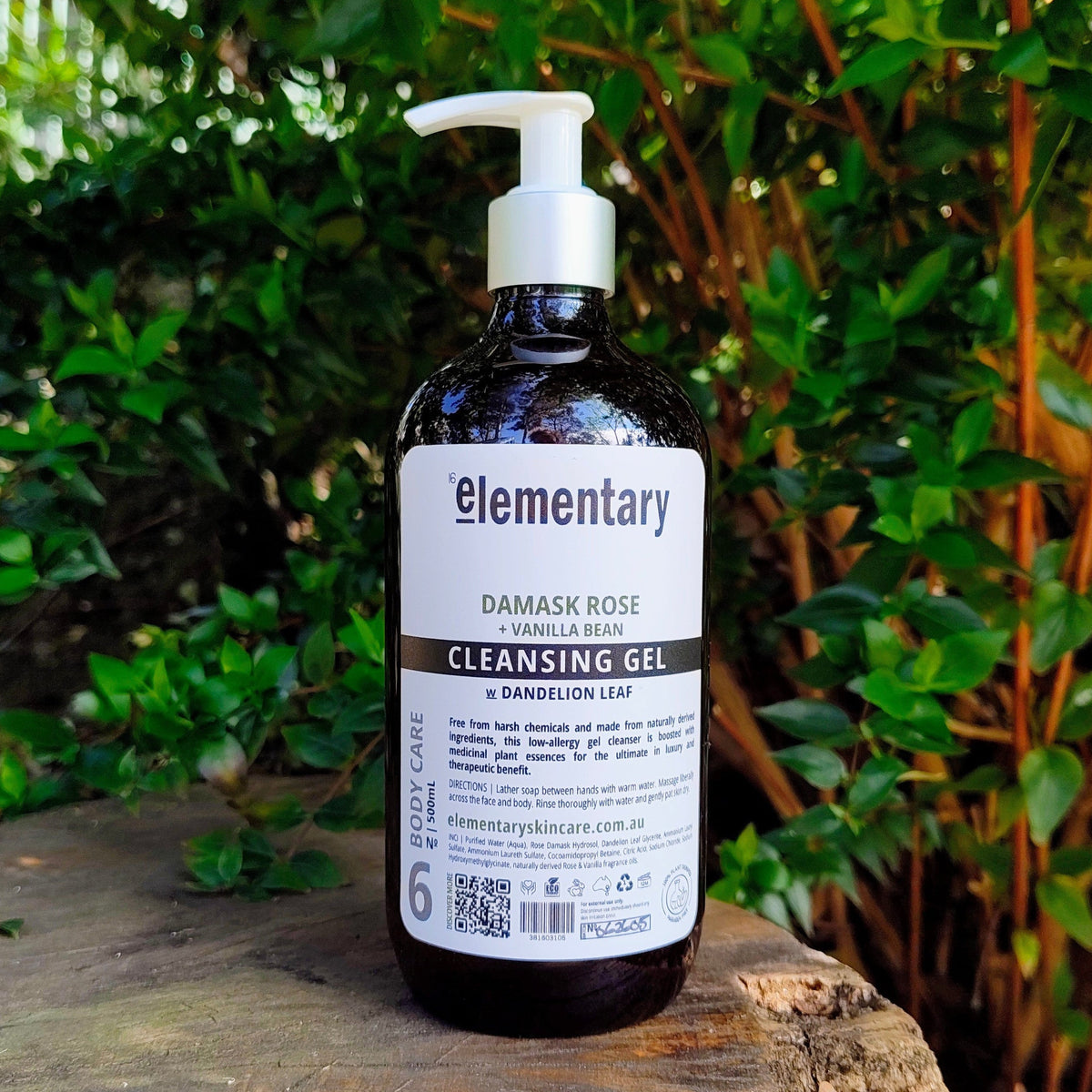 Elementary Damask Rose Cleansing Gel in a dark amber bottle with a white pump, placed on a rustic wooden log with a lush green garden background.