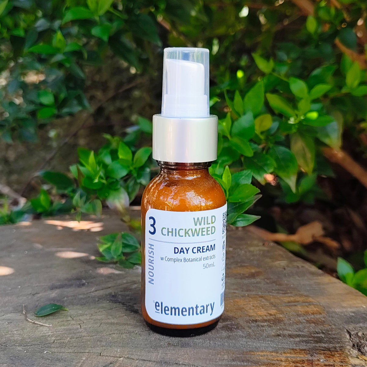 A small amber glass spray bottle of elementary brand Wild Chickweed Day Cream sits on a weathered wooden log with a blurred background of green foliage.