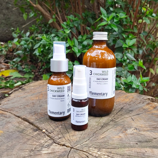 Three amber glass skincare bottles with white labels from the brand Elementary, including a large serum bottle and two smaller cream bottles, arranged on a weathered wooden log with green foliage in the background.