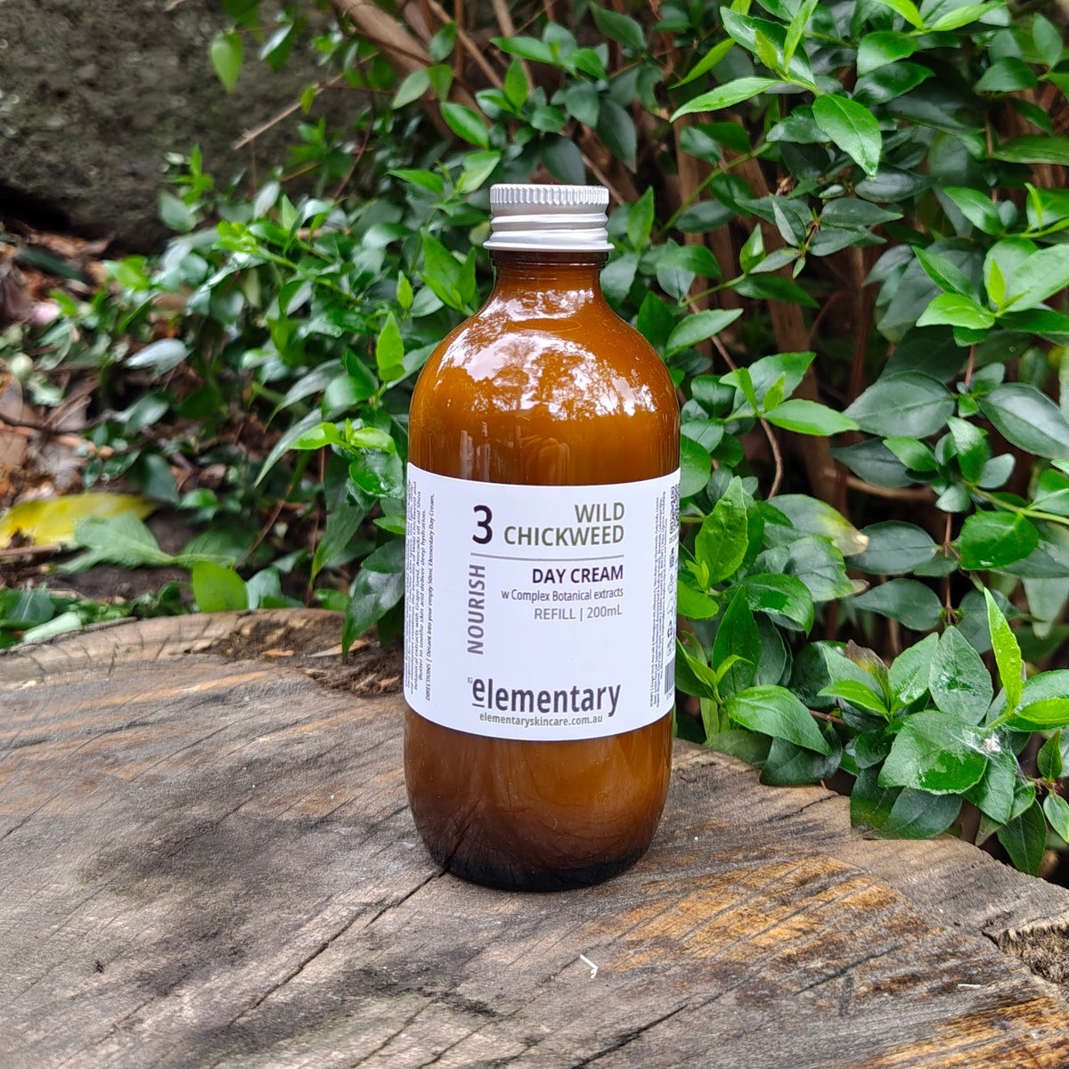 An 200ml Refill amber glass bottle of Elementary Wild Chickweed Day Cream sits on a weathered wooden log with lush green foliage in the background.